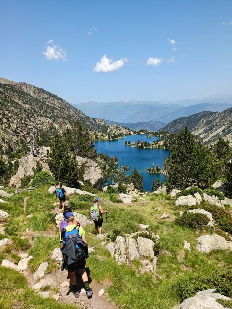 Un grup de senderistes amb motxilles caminant per un sender de muntanya vora un estany blau cap al refugi Josep Maria Blanch.