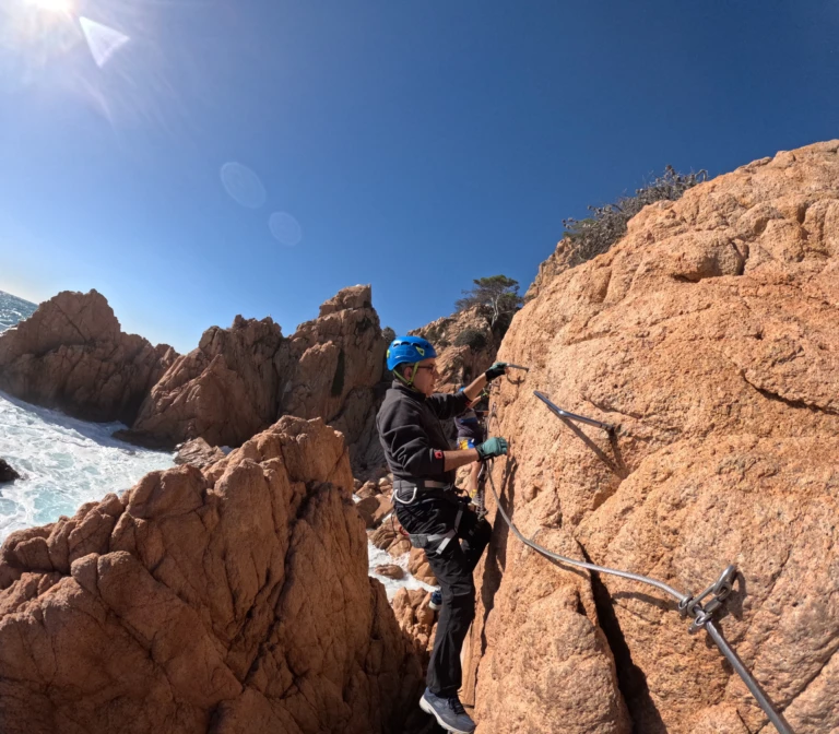Un home progressant per una paret de roca vertical equipada amb graons de ferro just a sobre del mar blau de la Costa Brava.